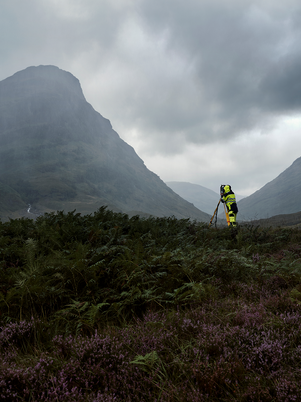 Person in high-visibility jacket standing in a mountainous landscape with misty mountains and overcast sky.